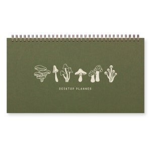 May include: A green desktop planner with a spiral binding. The cover features a line drawing of various mushrooms in white, with the words "DESKTOP PLANNER" below the illustrations. The planner is rectangular and sits on a white background.