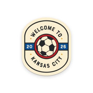 May include: A cream-colored sticker with a soccer ball graphic inside a red circle. The text "WELCOME TO KANSAS CITY 2026" is printed around the soccer ball in a black, bold font. The sticker has a rounded rectangular shape.
