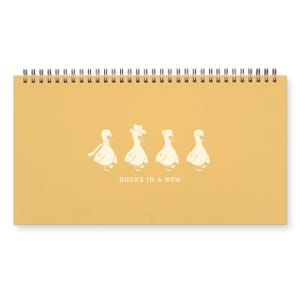 May include: A spiral-bound notepad with a mustard-yellow cover. The cover features four cartoon ducks, each wearing a different accessory, with the text "DUCKS IN A ROW" below them. The notepad is designed for writing and note-taking.