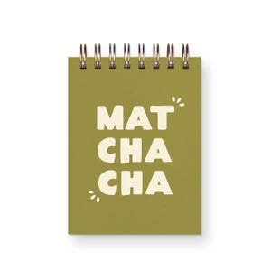 May include: A spiral-bound notebook with a matte olive green cover. The front cover features the text MAT CHA CHA in bold, cream-colored, rounded capital letters, accompanied by small decorative leaf-like accents above and below the text.