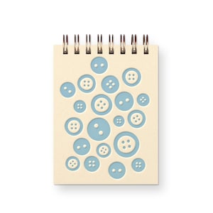 May include: A spiral-bound notebook with a beige cover featuring a pattern of light blue buttons in various sizes. The buttons have white centers and are arranged in a scattered design. The spiral binding is gold-colored.