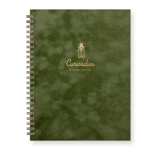 The Curiosities journal in a suede textured moss green features a gold foil cicada illustration and the words "Curiosities and other oddities". Journal is spiral bound on the side.