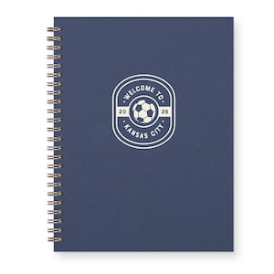 May include: A navy blue spiral-bound notebook. The cover features a white circular emblem with the text "WELCOME TO" above a soccer ball graphic, the numbers "20" and "26", and "KANSAS CITY" below. The spiral binding is gold.