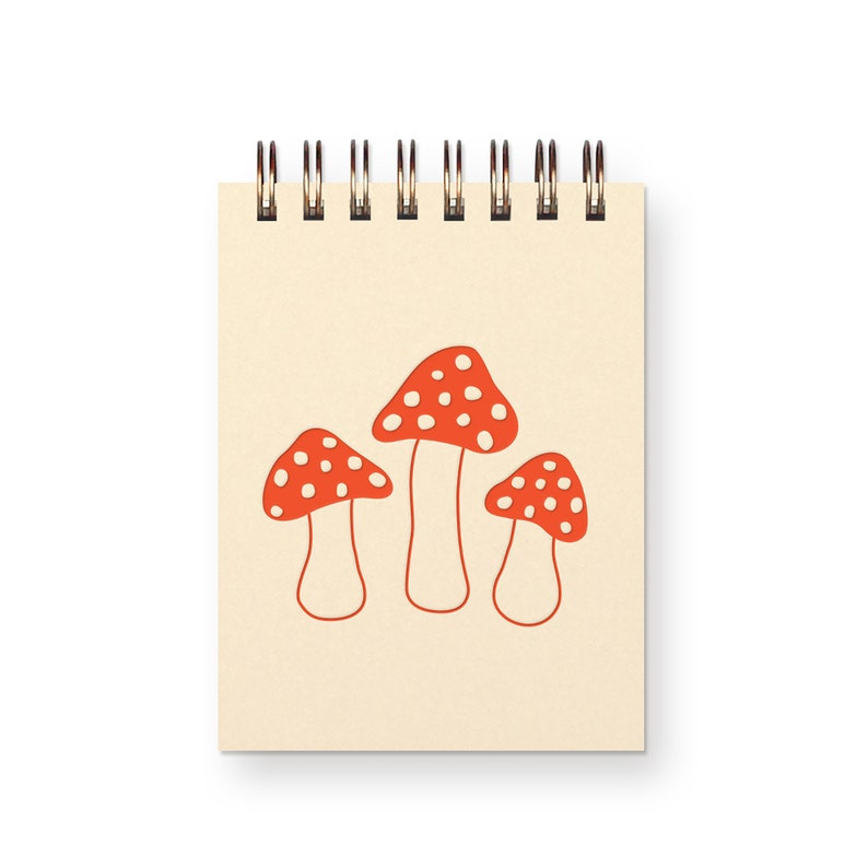 A small, top spiral bound ivory notebook with three red toadstool mushrooms on it.