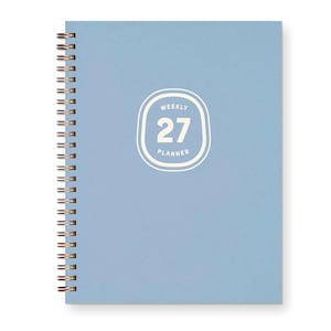 May include: A light blue spiral-bound weekly planner with a copper-colored metal coil on the left side. The front cover features a white graphic with rounded corners containing the text "WEEKLY 27 PLANNER" printed in a clean, sans-serif font.