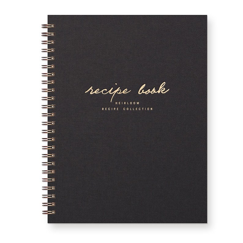 Spiral bound keepsake recipe book with black linen cover. Gold foil font reads "recipe book heirloom recipe collection."