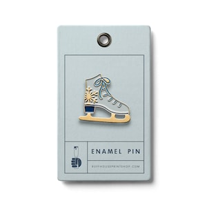 May include: White enamel pin of an ice skate with a gold snowflake and blue laces. The pin is on a light blue card with the text "ENAMEL PIN" and "RUFFHOUSEPRINTSHOP.COM".