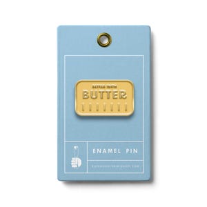 May include: A gold-colored enamel pin in the shape of a butter stick, with the words "BETTER WITH BUTTER" embossed on it. The pin is displayed on a light blue card with the words "ENAMEL PIN" and a website address.