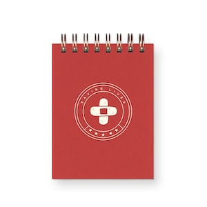 May include: A red spiral-bound notepad with a white and beige circular logo featuring a bandage cross and the words "SAVING LIVES". The notepad has a clean, minimalist design.