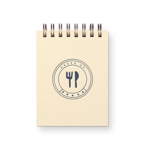 May include: A small notepad with a spiral binding and a cream-coloured cover. The cover features a blue circular design with a fork and knife, and the words "ORDER UP".