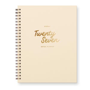 May include: A cream-colored spiral-bound weekly dated planner with gold foil lettering on the front cover that reads "WEEKLY Twenty Seven DATED PLANNER". The planner features a metallic bronze wire binding along the left side of the cover.