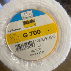 May include: A roll of white fabric in clear wrapping. The label is yellow and blue, with the text "Vlieseline G 700". The label also states the fabric is 90 cm x 25 m and 100% CO.