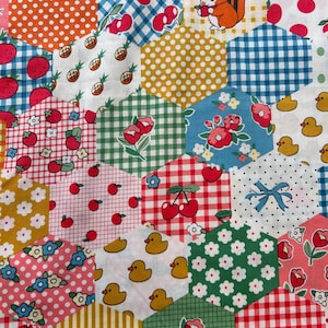May include: A colourful fabric with a patchwork design featuring hexagonal shapes. Each hexagon has a different pattern, including pineapples, cherries, strawberries, ducks, flowers, and gingham.