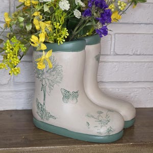 May include: A pair of cream-colored boot-shaped vases with a green trim and floral and butterfly designs. The vases are filled with yellow, white, and purple flowers, creating a decorative display.