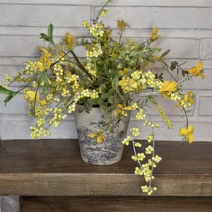 May include: A decorative floral arrangement in a ceramic pot. The pot features a blue and white toile design with butterflies. The arrangement includes yellow flowers and berries, with green foliage. The pot sits on a wooden shelf.