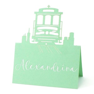 May include: A light green paper place card featuring a cut-out design of a San Francisco cable car and skyline. The name "Alexandrina" is written in cursive script on the card.