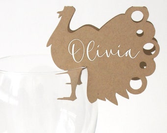 May include: A clear wine glass with a brown turkey-shaped place card. The place card has the name "Olivia" written in white script. The turkey has a decorative tail and is perched on the rim of the glass.