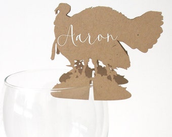 Puede incluir: Una tarjeta de mesa con forma de pavo marrón con el nombre "Aaron" en escritura cursiva, apoyada en el borde de una copa de vino transparente. La tarjeta está hecha de un material marrón claro, sobre fondo blanco.