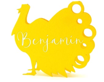 May include: A yellow turkey-shaped place card with the name "Benjamin" in a cursive font. The turkey has a detailed tail and is set against a white background. Ideal for Thanksgiving or fall-themed events.