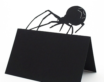May include: A black paper place card with a die-cut spider silhouette on top. The spider has long legs and a rounded body. The place card is folded in half, creating a tent shape, and is set against a white background.