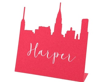 May include: A red cardstock place card featuring a city skyline silhouette. The name "Harper" is written in white script across the front. The card is folded at the bottom to stand upright.
