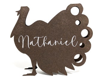 Puede incluir: Una tarjeta de lugar de madera marrón con forma de pavo con el nombre "Nathaniel" en escritura blanca. El pavo tiene una cola detallada con recortes circulares. Ideal para Acción de Gracias.