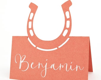 May include: Coral-colored place card with a horseshoe cutout at the top. The name "Benjamin" is written in white script across the front. The card is folded to stand upright.