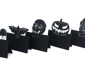 May include: Black Halloween-themed place card holders featuring a spider, bat, skull, jack-o'-lantern, and zombie. Each card holder has a cutout design on top of a black rectangular base, perfect for a spooky party.