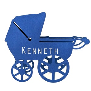 Op de afbeelding: Een blauwe papieren kinderwagen met de naam "KENNETH" in witte letters. De wagen heeft een kap, vier wielen en een handvat. Het ontwerp is uit het papier gesneden.