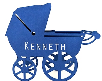 May include: A blue paper baby carriage with the name "KENNETH" in white lettering. The carriage has a hood, four wheels, and a handle. The design is cut out of the paper.