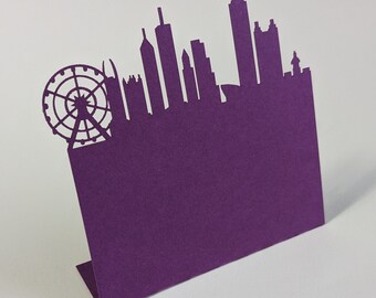 May include: A purple paper card featuring a city skyline silhouette. The design includes a Ferris wheel and various building shapes. The card is folded to stand upright, showcasing the detailed cut-out design.