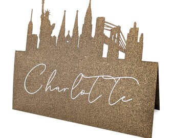 May include: A gold glitter cardstock place card featuring a city skyline silhouette. The skyline includes the Statue of Liberty and other iconic buildings. The name "Charlotte" is written in white script across the front.
