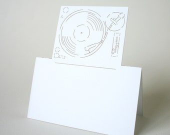 May include: A white card with a laser-cut design of a turntable. The card is folded, with the turntable design at the top. The turntable design includes the record, tone arm, and other details. The card is blank, ready for a message.