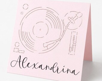 May include: A pink card with a cut-out design of a record player. The name "Alexandrina" is written in cursive script below the record player design. The card is standing upright on a white surface.