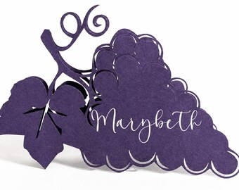 May include: A deep purple paper place card shaped like a cluster of grapes with a vine and leaf. The name Marybeth is printed in white cursive script across the center of the grape cluster. The design features die-cut edges and a textured finish.