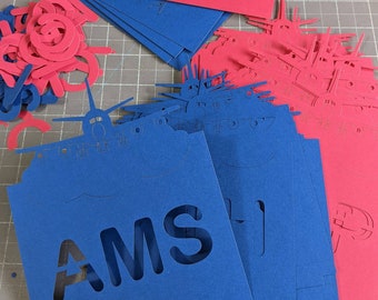 May include: A collection of blue and red paper cutouts. Blue cutouts feature the letters "AMS" and ship designs. Red cutouts also feature ship designs. The cutouts are arranged on a gray cutting mat.