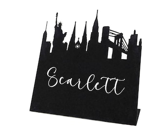 May include: A black place card featuring a silhouette of the New York City skyline, including the Statue of Liberty and the Brooklyn Bridge. The name "Scarlett" is written in white script across the front.