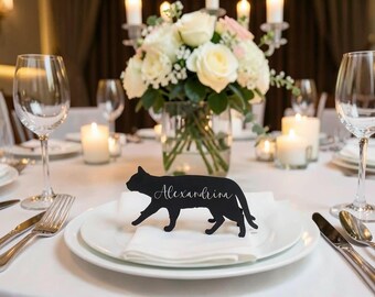 May include: A black cat-shaped place card with the name "Alexandrina" in cursive script, placed on a white napkin atop a white plate. The table setting includes silverware, wine glasses, and a floral centerpiece with candles.