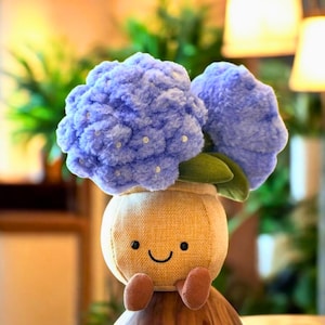 May include: A plush toy shaped like a flower with a light blue, textured bloom. The flower sits atop a tan pot with a smiling face and small brown feet. Green leaves peek out from under the flower.