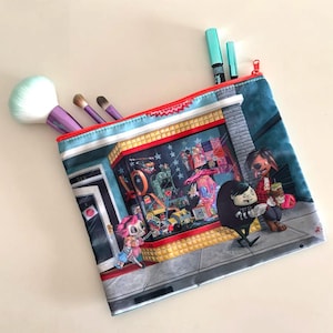 May include: A rectangular makeup bag with a colorful illustration of a storefront scene. The bag has a red zipper and contains makeup brushes and pens. The storefront features cartoon characters and toys.