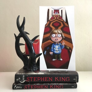 May include: A print of a cartoon child in a blue sweater, standing on a red and brown patterned carpet, with a hallway in the background. The print is propped up against two Stephen King books and a black decorative candle holder.