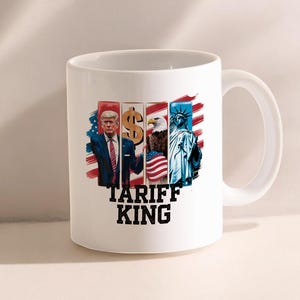 Puede incluir: Taza de café de cerámica blanca con un gráfico colorido de un hombre, un signo de dólar, un águila y la Estatua de la Libertad. El texto "Tariff King" está impreso debajo del gráfico.
