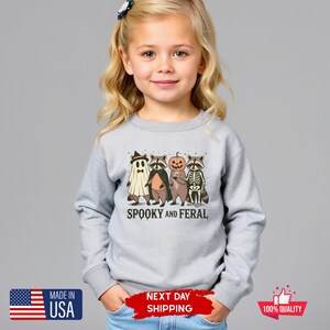 Puede incluir: Sudadera gris claro con un dise&ntilde;o de Halloween. El dise&ntilde;o presenta un fantasma, un mapache, una calabaza y un mapache esqueleto, con el texto "Spooky and Feral". Fabricado en los Estados Unidos.