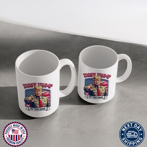 May include: Two white ceramic mugs with a graphic of Donald Trump on them. The graphic says "Tariff Trump" and "Pay Your Share". The mugs are on a white surface.