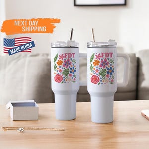 May include: Two white stainless steel travel mugs with lids and straws. Both mugs feature a floral design with pink, orange, blue, and green flowers and the text "FDT" in pink letters.