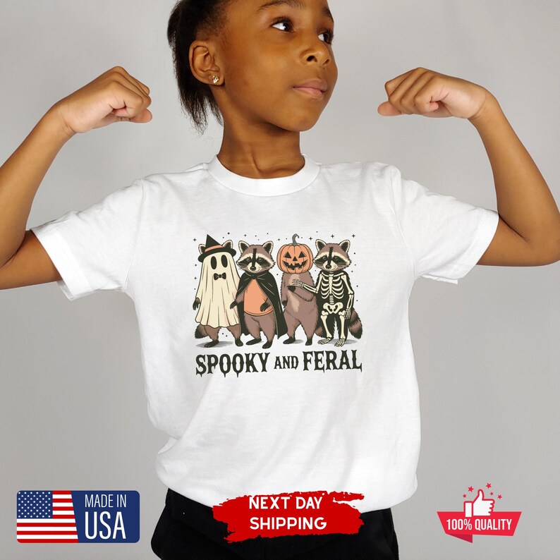 Puede incluir: Camiseta blanca con un dise&ntilde;o de Halloween. El gr&aacute;fico muestra cuatro mapaches disfrazados: un fantasma, un vampiro, una calabaza y un esqueleto. El texto "SPOOKY AND FERAL" est&aacute; impreso debajo. La camiseta tambi&eacute;n tiene una etiqueta "Made in USA".