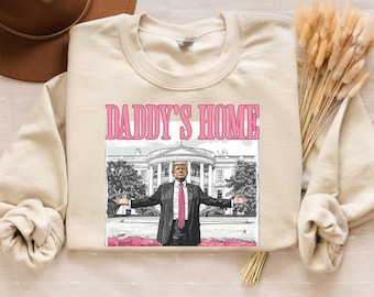 May include: A beige sweatshirt with a graphic print of a man in front of the White House. The text "Daddy's Home" is printed in pink above the image.