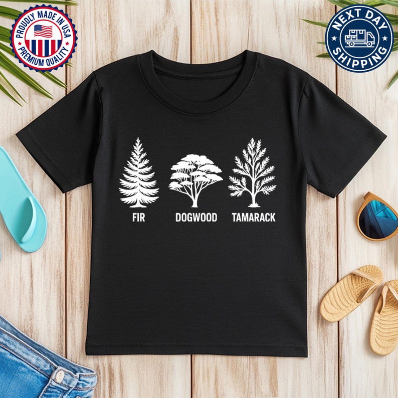 Pu&ograve; includere: T-shirt nera con tre illustrazioni di alberi bianchi e i nomi FIR, DOGWOOD e TAMARACK sotto. La maglietta ha un girocollo e maniche corte. L'immagine include anche il testo "MADE IN USA" e "NEXT DAY SHIPPING".