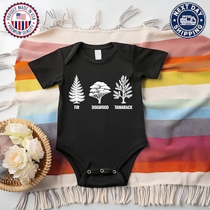Pu&ograve; includere: Body da bambino nero con illustrazioni di alberi bianchi e le parole "FIR", "DOGWOOD" e "TAMARACK" stampate sotto. Il body &egrave; su una coperta a righe colorata. Un cesto di fiori bianchi &egrave; visibile.