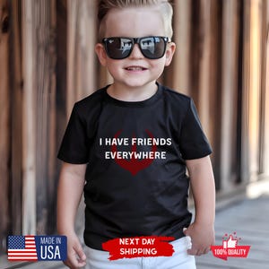 May include: A black t-shirt with the text "I HAVE FRIENDS EVERYWHERE" in white and a red graphic. The image shows a young person wearing sunglasses and the t-shirt. The shirt is made in the USA.